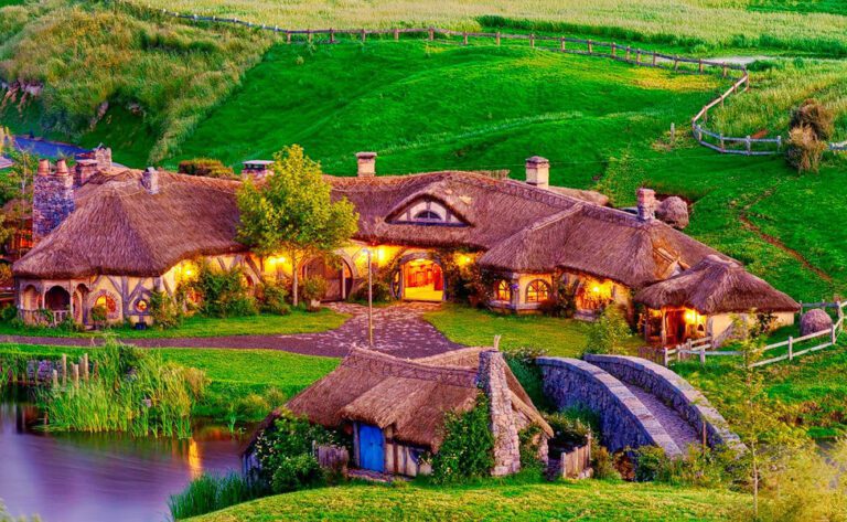 פונדק הדרקון הירוק (Green Dragon Inn) בהוביטון, ניו זילנד, עם גגות קש, אורות חמים המשתקפים באגם וגבעות ירוקות מסביב בשעת בין הערביים.