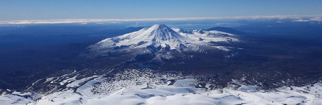 הנה התיאורים להר נגאורוהו (Mount Ngauruhoe), הלב הפועם והמושלג של הפארק הלאומי טונגרירו: Alt Text: מבט פנורמי מהאוויר על הר נגאורוהו (Mount Ngauruhoe) בניו זילנד, כשהוא מכוסה בשכבת שלג לבנה ועמוקה, על רקע שמיים כחולים צלולים ואגם טאופו המנצנץ באופק.