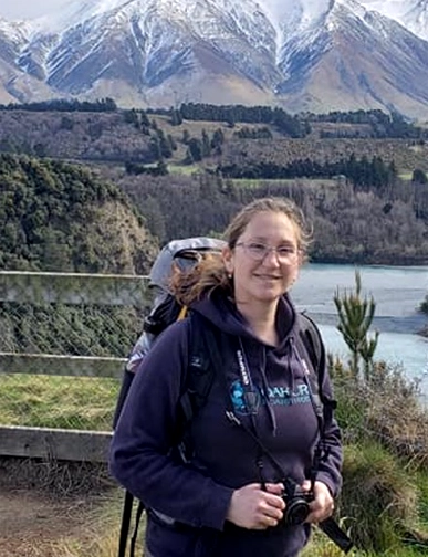 יפעת מצוות Rotem NZ מטיילת עם תרמיל ומצלמה מול אגם והרים מושלגים בניו זילנד