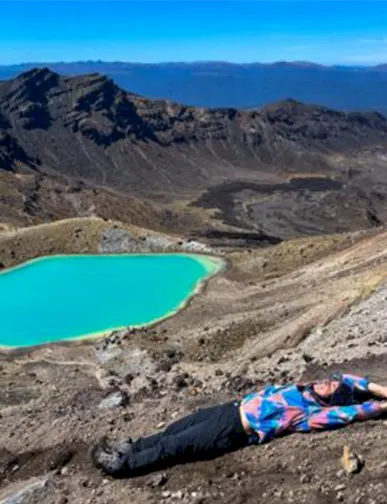 מטיילת שוכבת על מדרון הררי מול אגם טורקיז בלב נוף וולקני בניו זילנד
