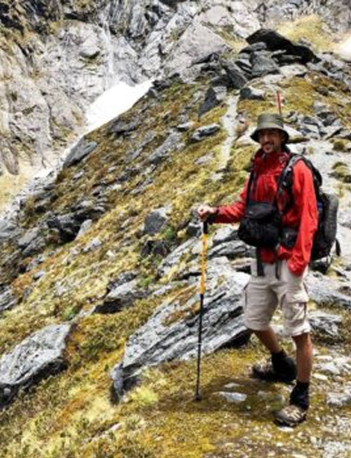 יונתן ברק עומד בשטח הררי עם מקל הליכה וציוד צילום